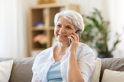 Eine Frau hat ein Telefon in der Hand. Sie befindet sich im Wohnzimmer. Sie lächelt.  | © Adobe Stock